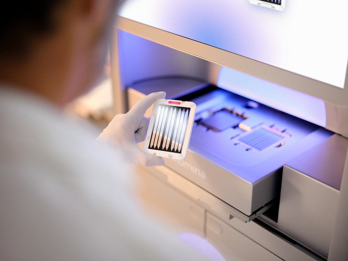 Close up and blurry image of the back side of a male scientist examining a second 25B flow cell cartridge before inserting into a NovaSeq X drawer.